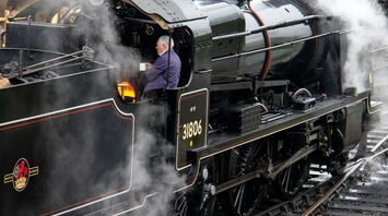A heritage steam locomotive in operation with a fireman tending to the firebox, surrounded by steam