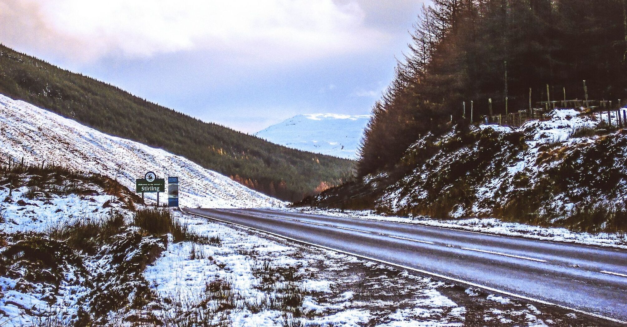 Snow-covered roadside in Scotland with a Stirling sign and distant hills under a cloudy sky