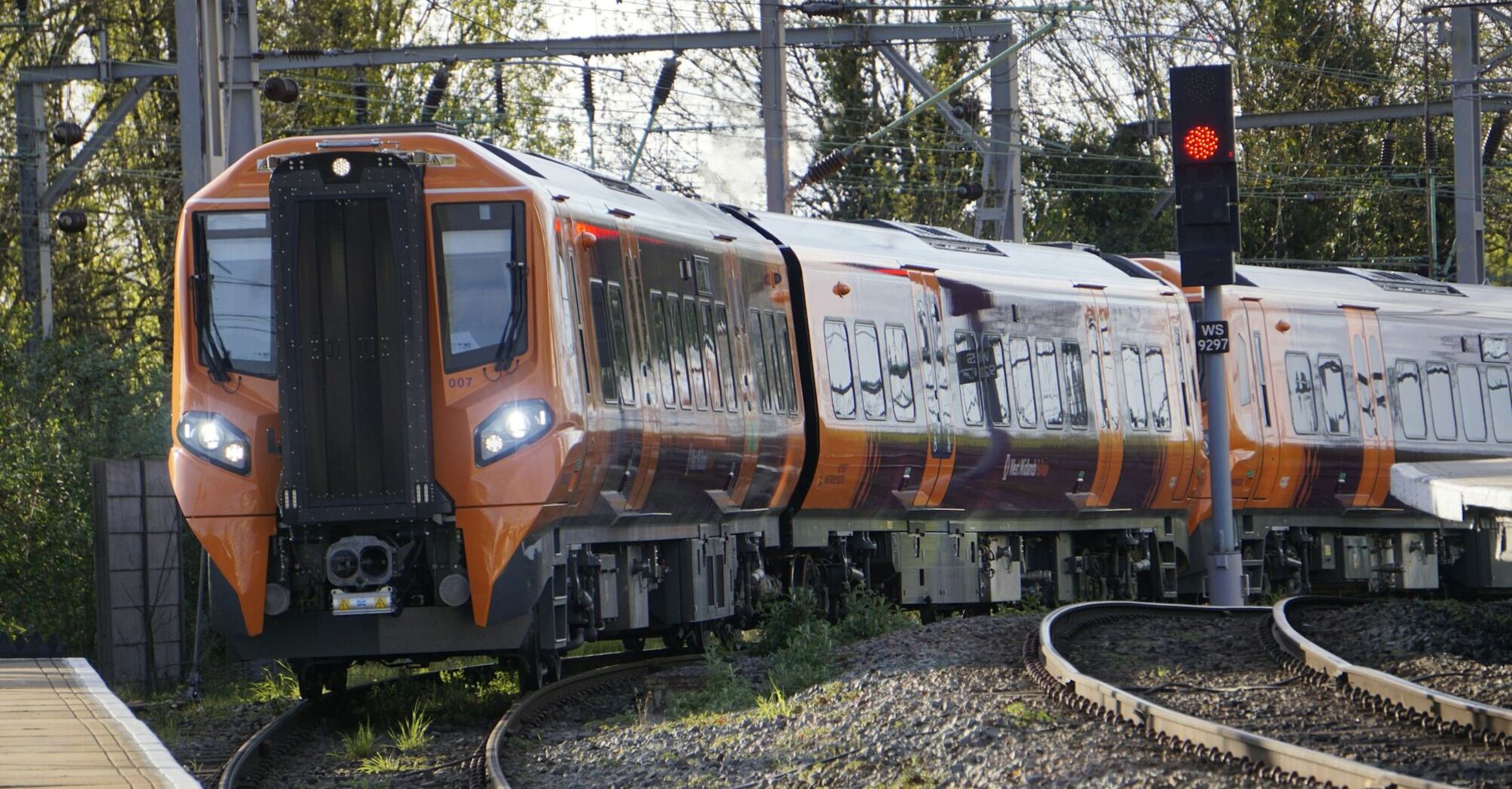 A bright orange West Midlands Railway train navigating a curved track near a platform, with trees and overhead electric lines in the background