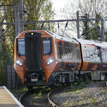 A bright orange West Midlands Railway train navigating a curved track near a platform, with trees and overhead electric lines in the background