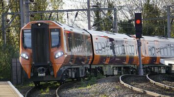A bright orange West Midlands Railway train navigating a curved track near a platform, with trees and overhead electric lines in the background
