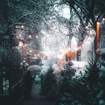 Snow-covered street with festive string lights, Christmas trees, and people holding umbrellas