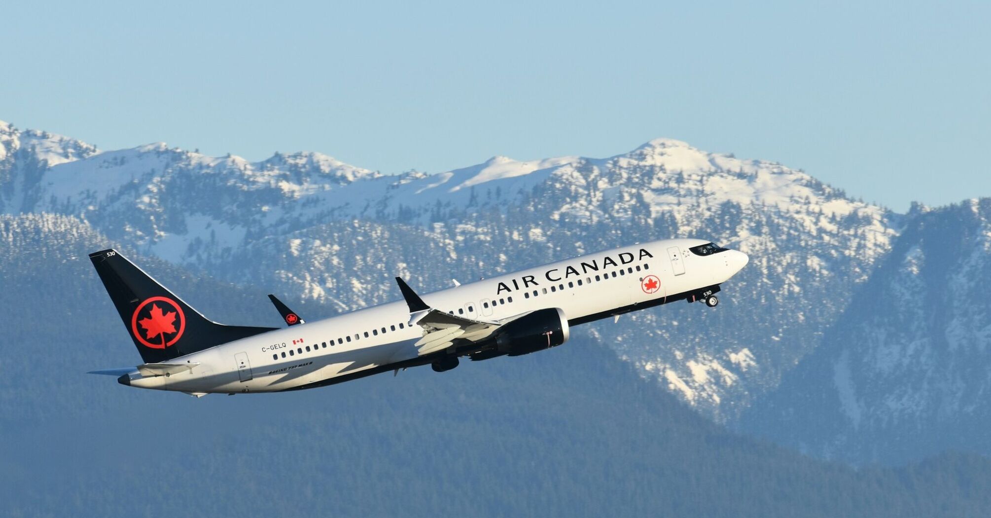 A white Air Canada plane takes off against a backdrop of snow-capped mountains under a clear blue sky