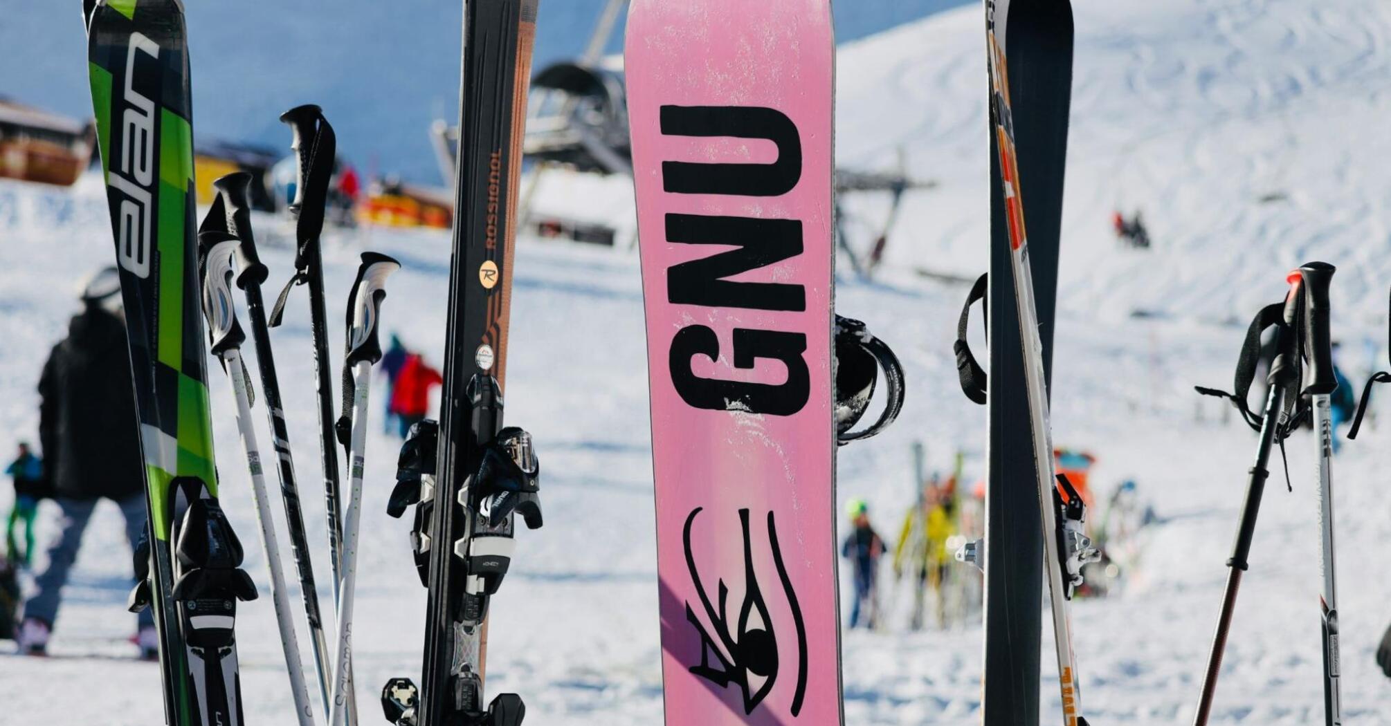 A collection of skis and snowboards on a snowy slope, with people and ski lifts in the background