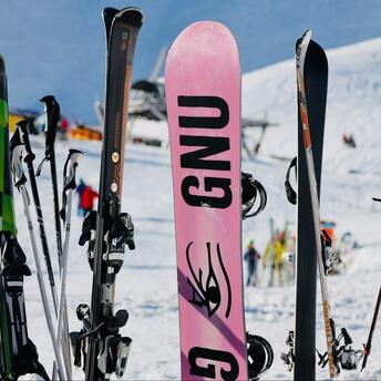 A collection of skis and snowboards on a snowy slope, with people and ski lifts in the background