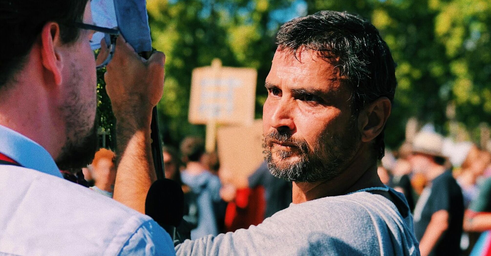 A man holding a sign on a protest with people and trees in the background