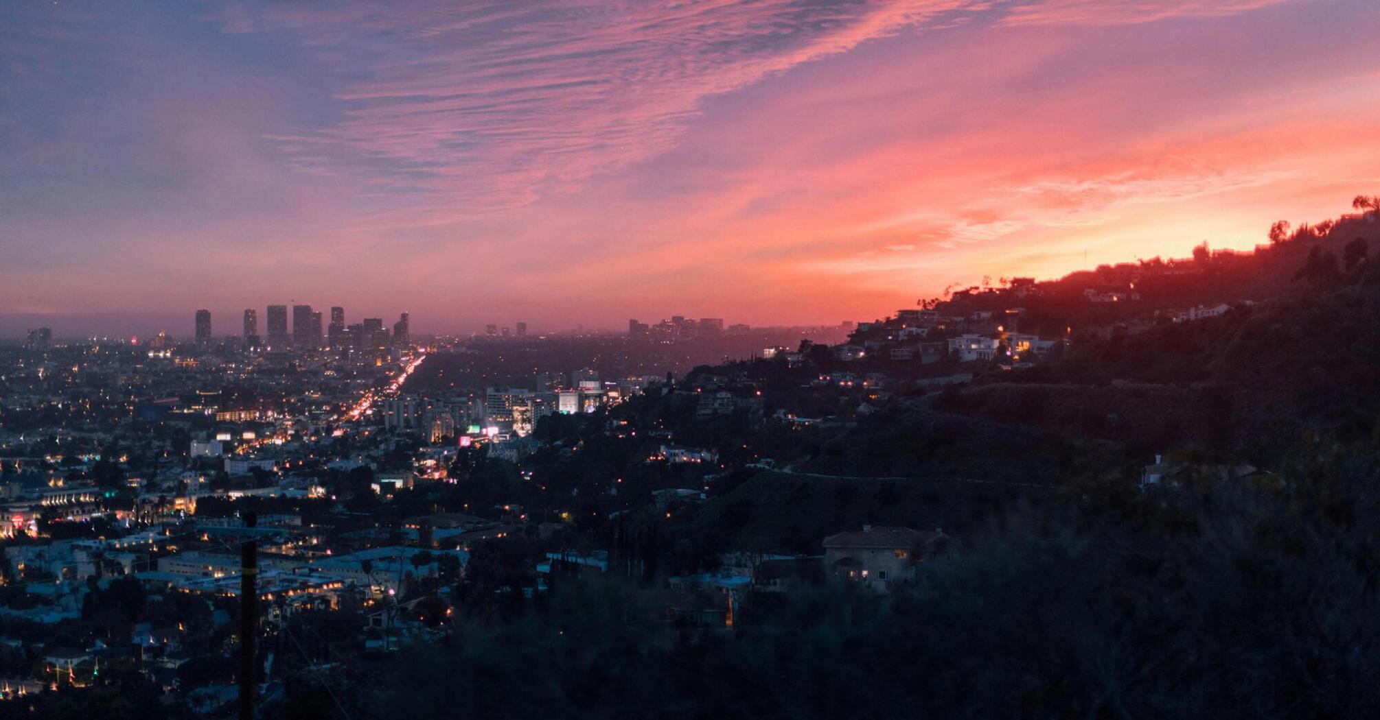 Sunset view of a cityscape with vibrant pink and orange skies, overlooking a densely lit urban area surrounded by hills