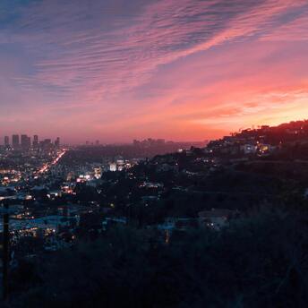 Sunset view of a cityscape with vibrant pink and orange skies, overlooking a densely lit urban area surrounded by hills