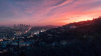 Sunset view of a cityscape with vibrant pink and orange skies, overlooking a densely lit urban area surrounded by hills