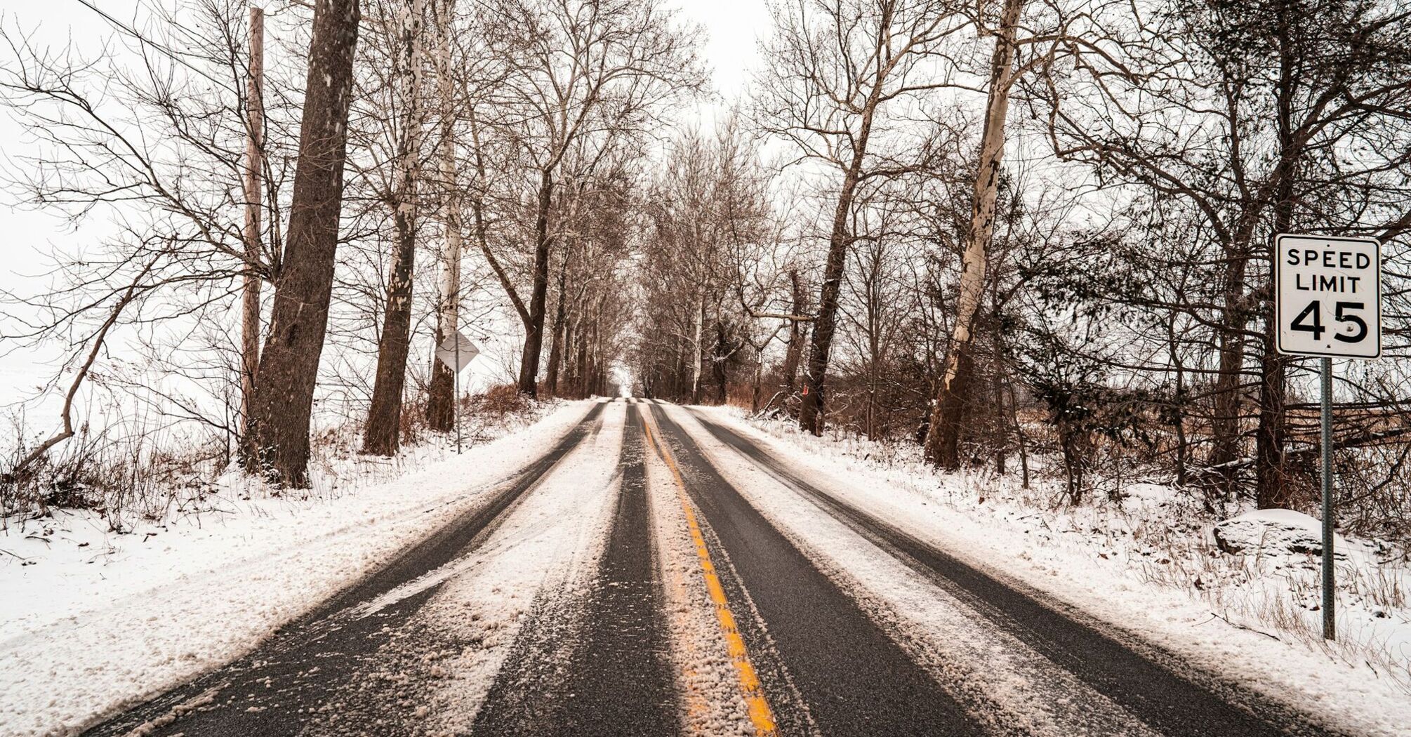 Snow-covered rural road with bare trees and a speed limit sign