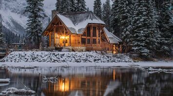 A cozy wooden cabin surrounded by snow-covered trees, reflected in a calm winter lake