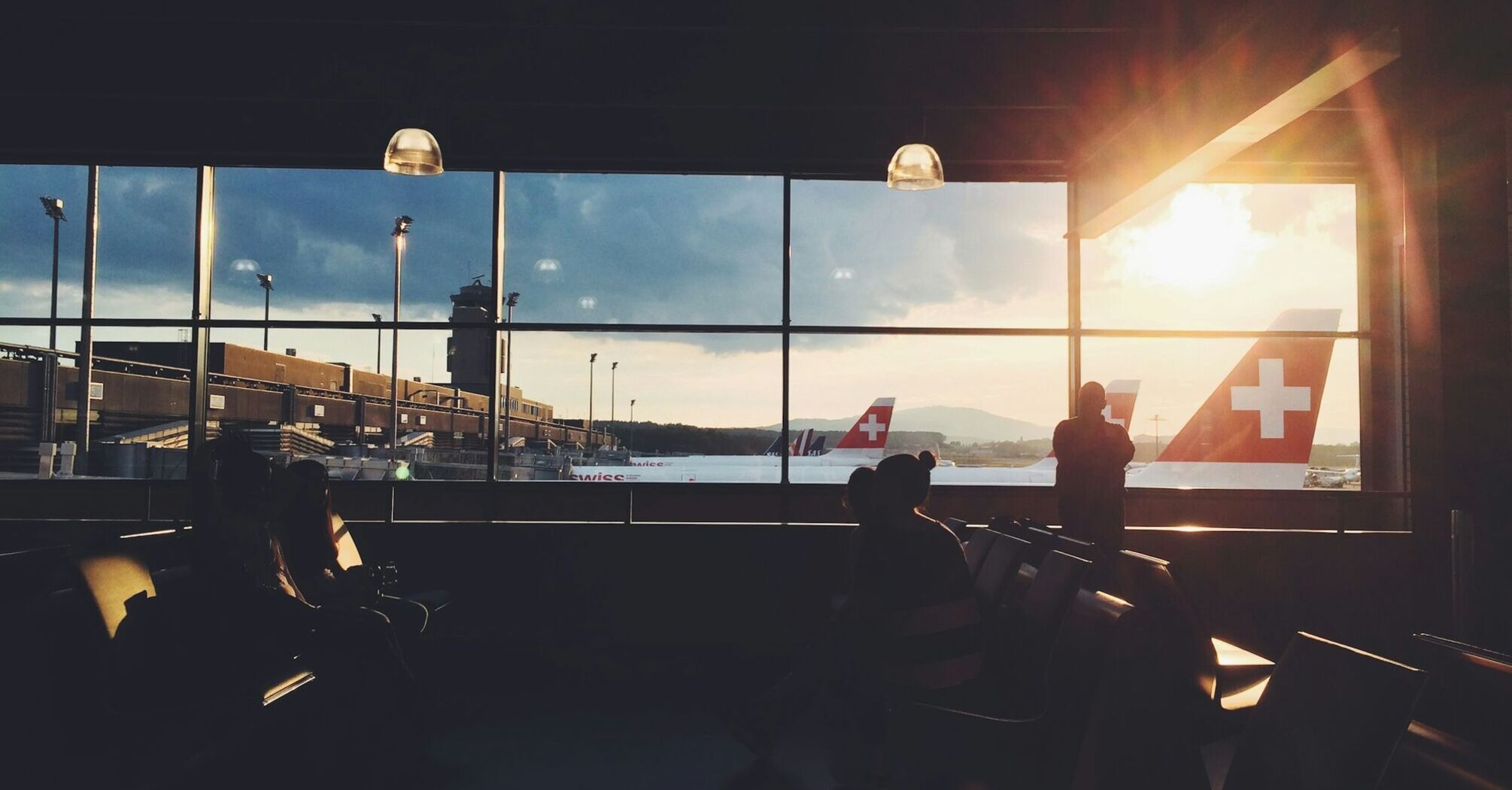 Travelers waiting inside an airport terminal at sunset with Swiss Air planes visible through the window