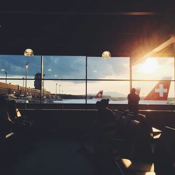 Travelers waiting inside an airport terminal at sunset with Swiss Air planes visible through the window