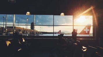 Travelers waiting inside an airport terminal at sunset with Swiss Air planes visible through the window