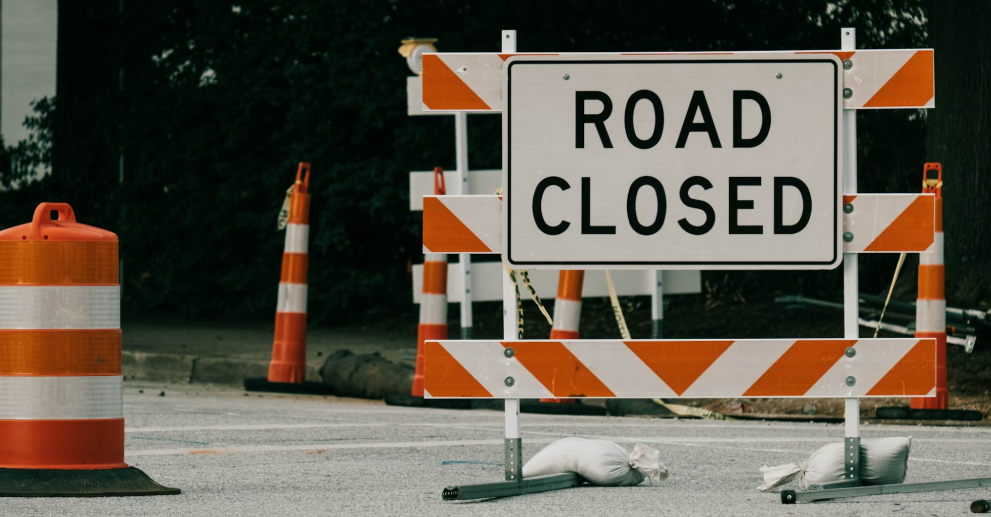 A road closed sign with orange traffic barrels and barricades indicating a blocked roadway