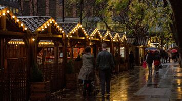 A rainy evening at Manchester Christmas Markets, with wooden stalls glowing under string lights and visitors strolling along the pathway