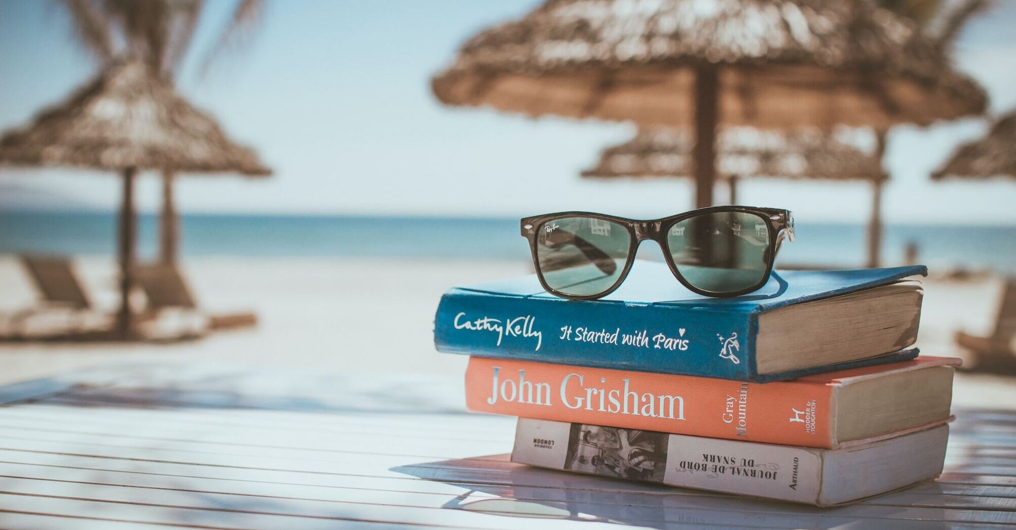 A stack of books and sunglasses against the backdrop of beach umbrellas and sun loungers