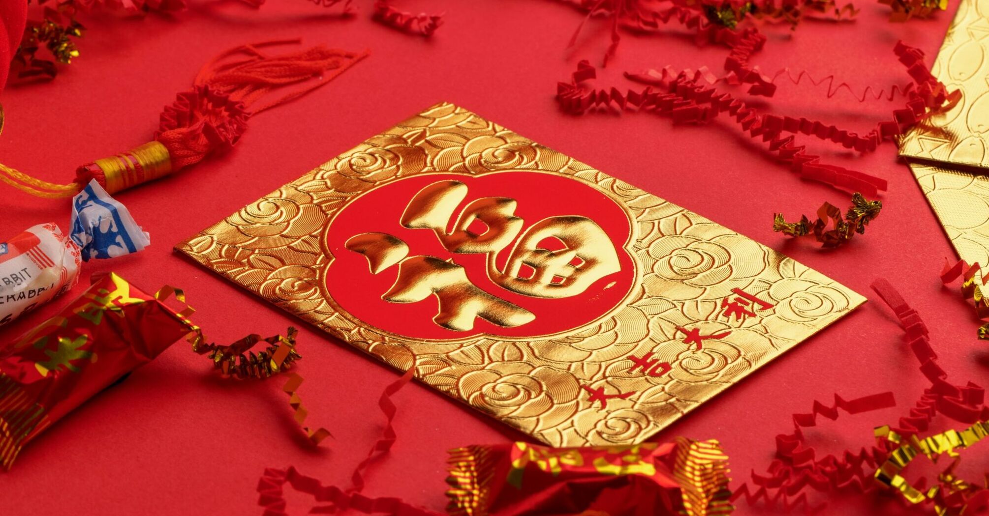 Traditional red envelopes adorned with gold Chinese characters, surrounded by red and gold festive decorations and firecracker replicas on a red background