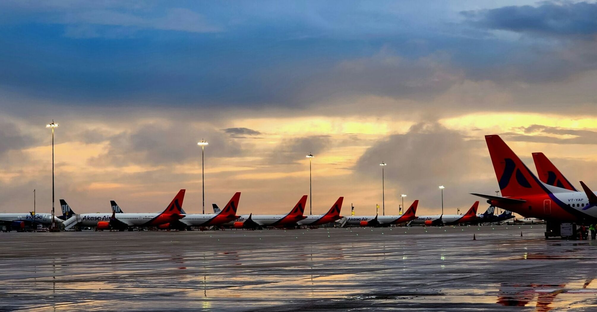 Line of Akasa Air planes on a wet tarmac at dusk with lit lampposts and a dramatic cloudy sky in the background