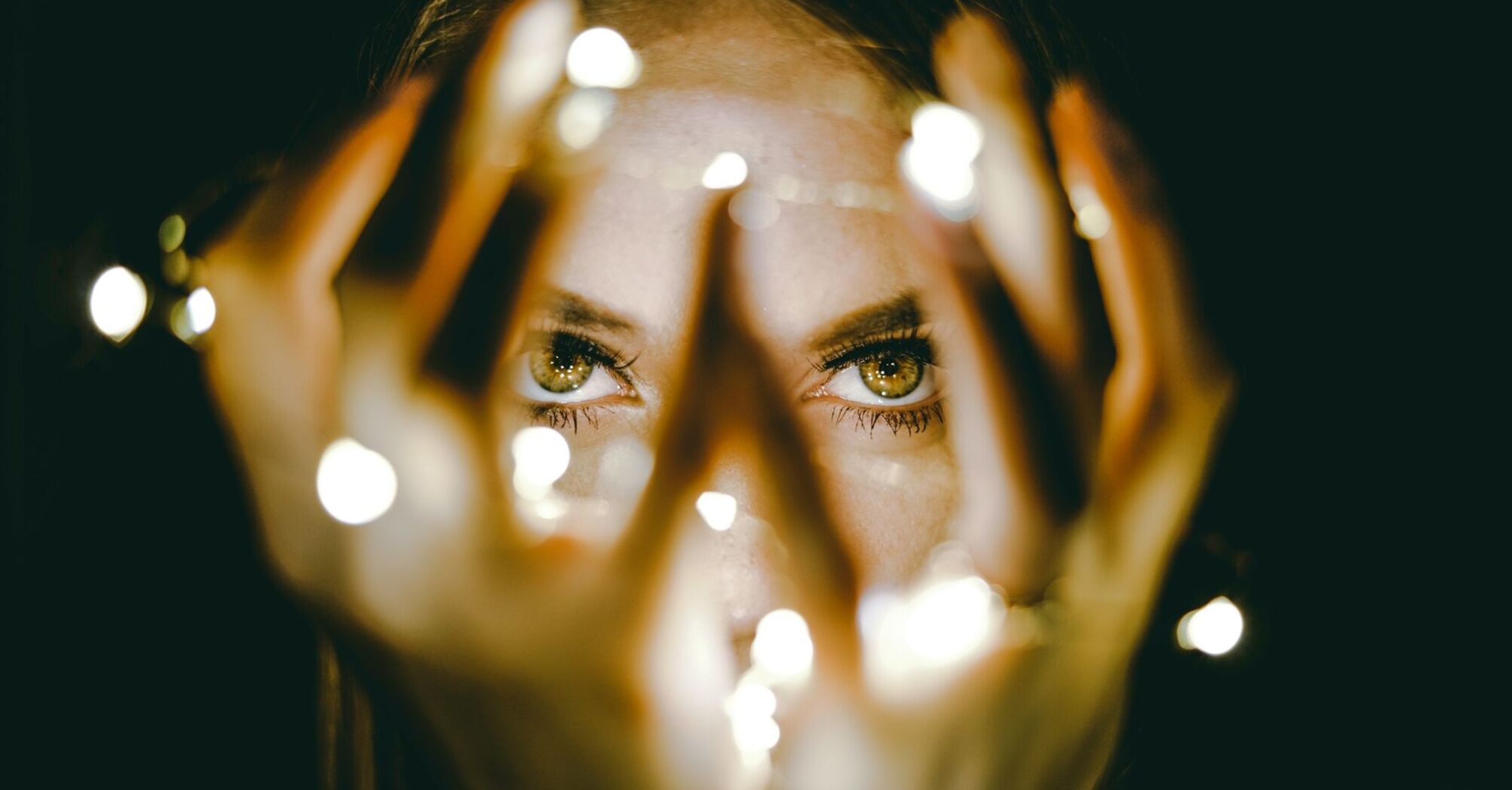 A woman's eyes gazing between her fingers, with soft lights creating a bokeh effect around her hands