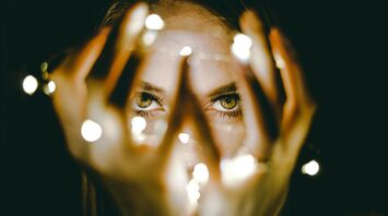 A woman's eyes gazing between her fingers, with soft lights creating a bokeh effect around her hands