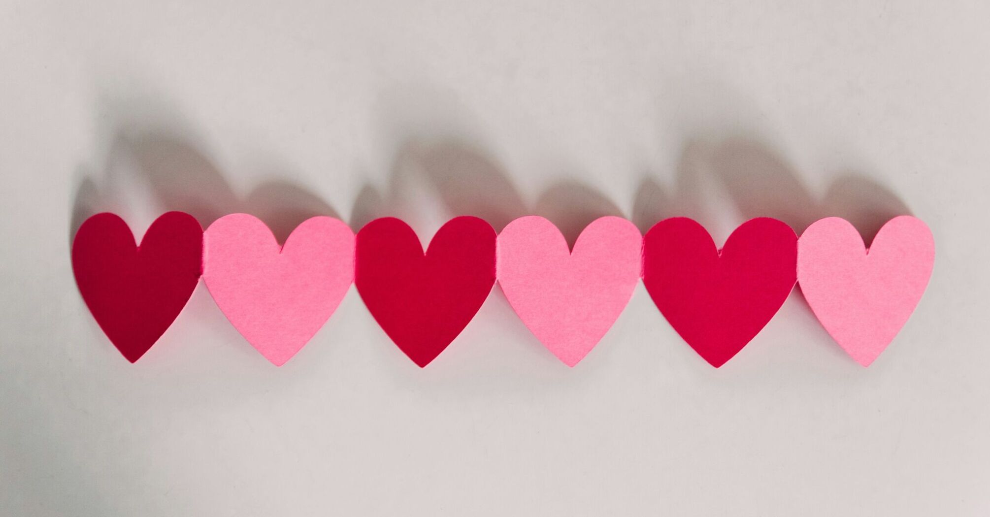A row of cut-out paper hearts in shades of pink and red with their shadows cast on a light background