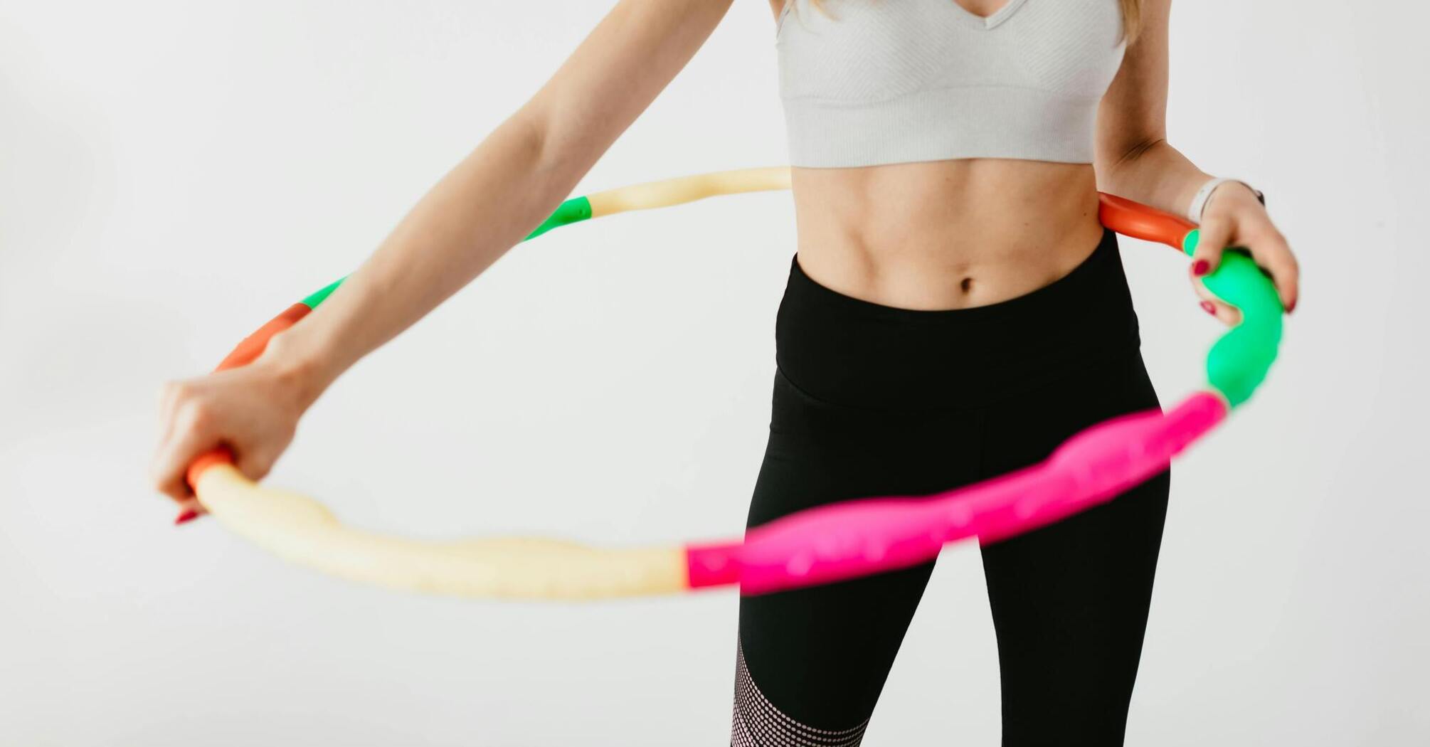 A person in a white sports top and black leggings holding a colorful hula hoop at waist level