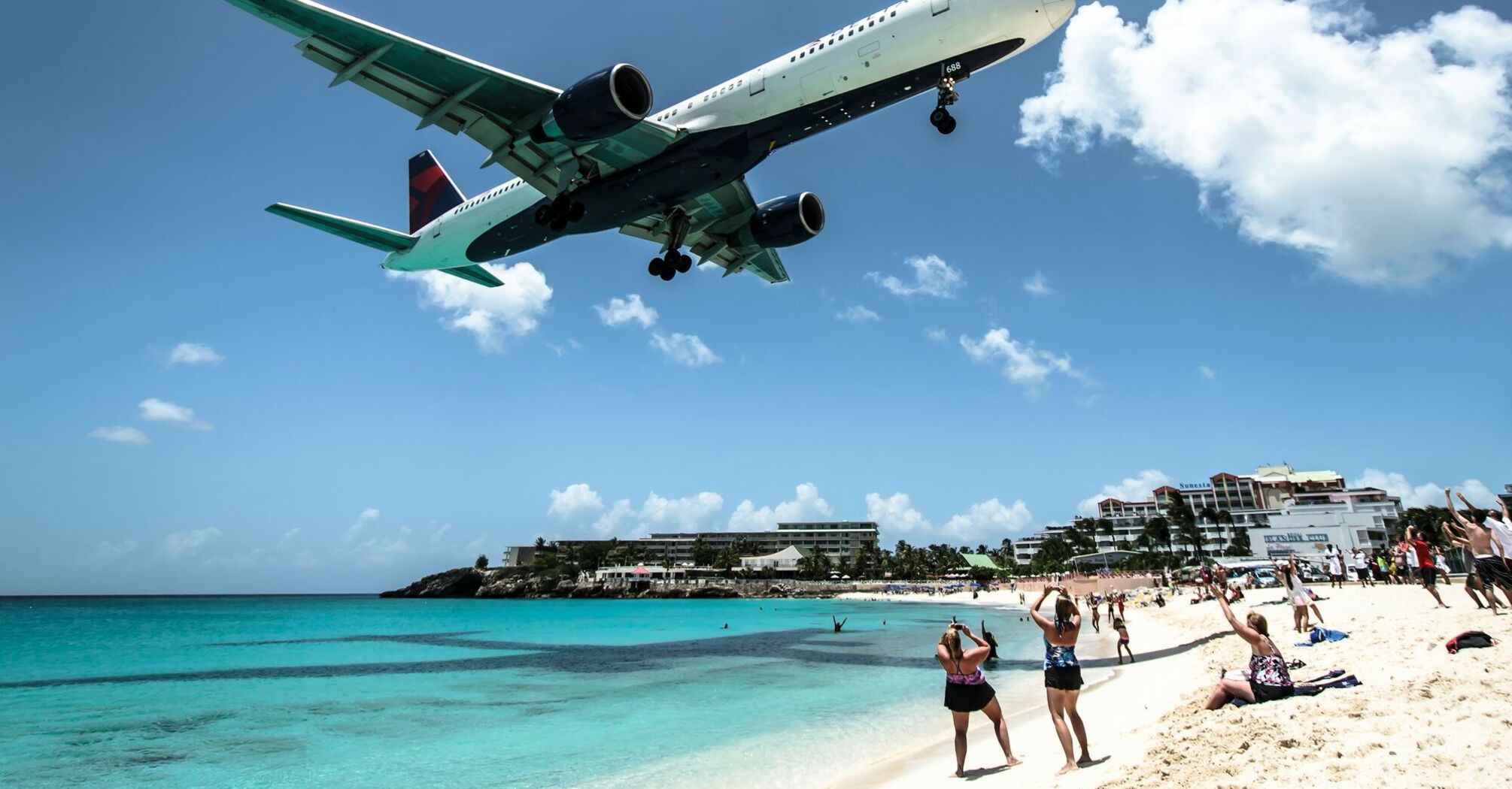 A Delta airplane landing over a sunny beach with clear turquoise waters, as onlookers on the sand take photos and watch 