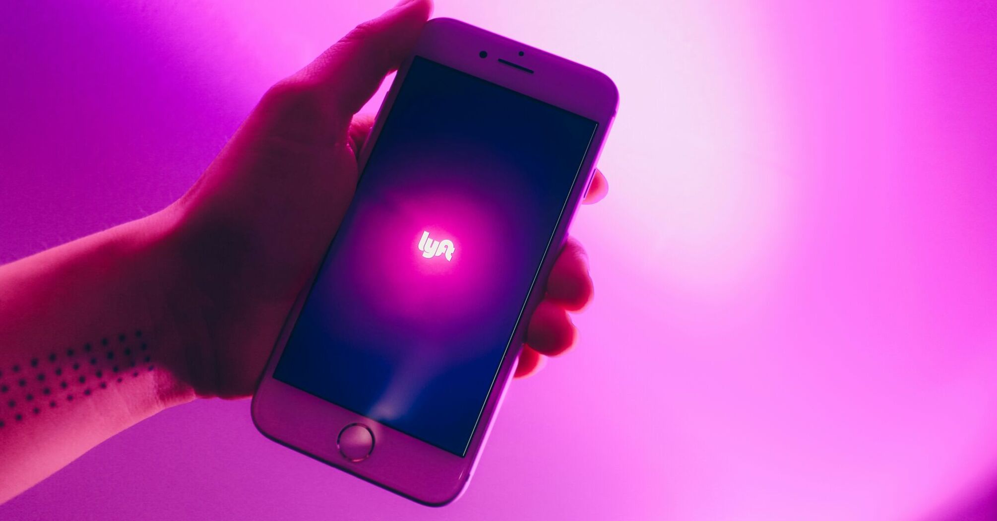Hand holding a smartphone with the Lyft app logo illuminated on the screen, set against a vibrant pink backdrop