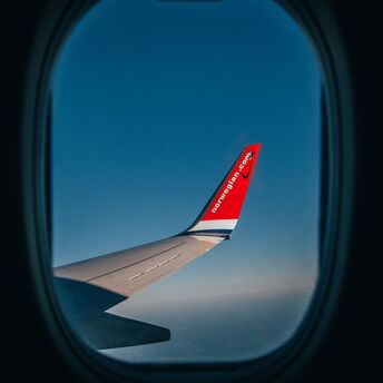 View from an airplane window showing the wing with Norwegian Air's logo against a clear blue sky