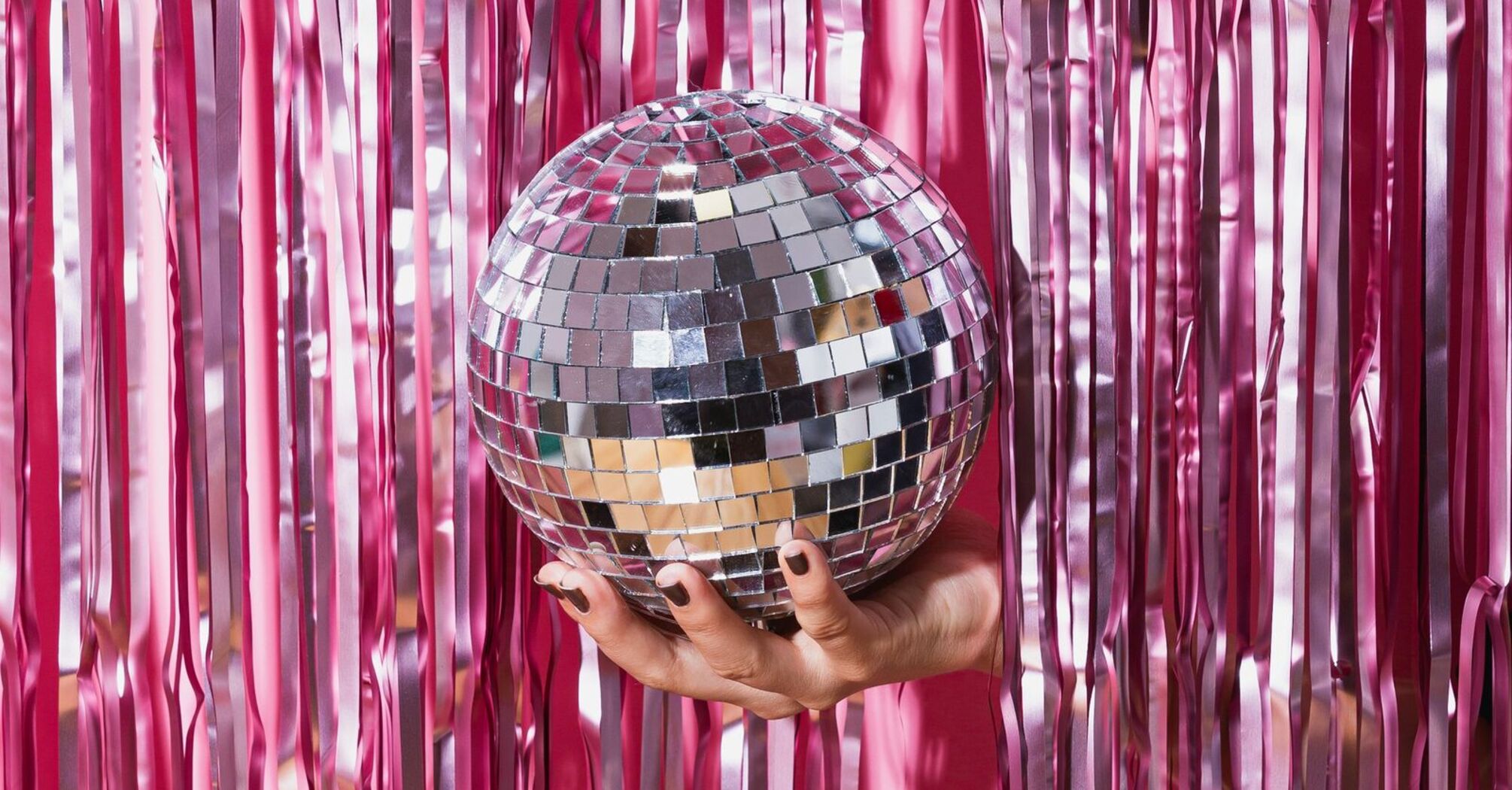 A person holding a disco ball in front of a background of shiny pink tinsel streamers