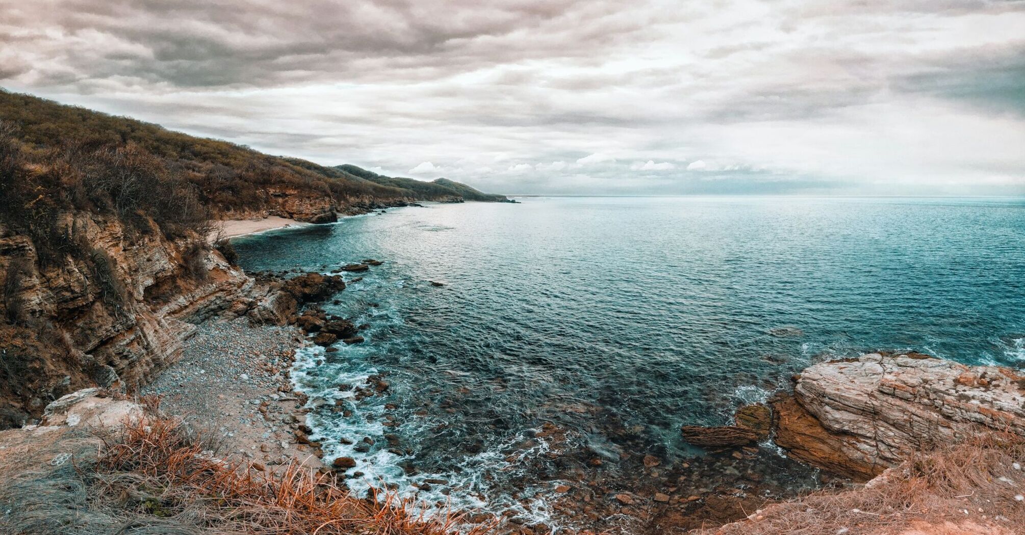 A serene coastal landscape of Nayarit, Mexico, featuring a pebbled beach, clear blue waters, and a rugged cliff under a cloudy sky