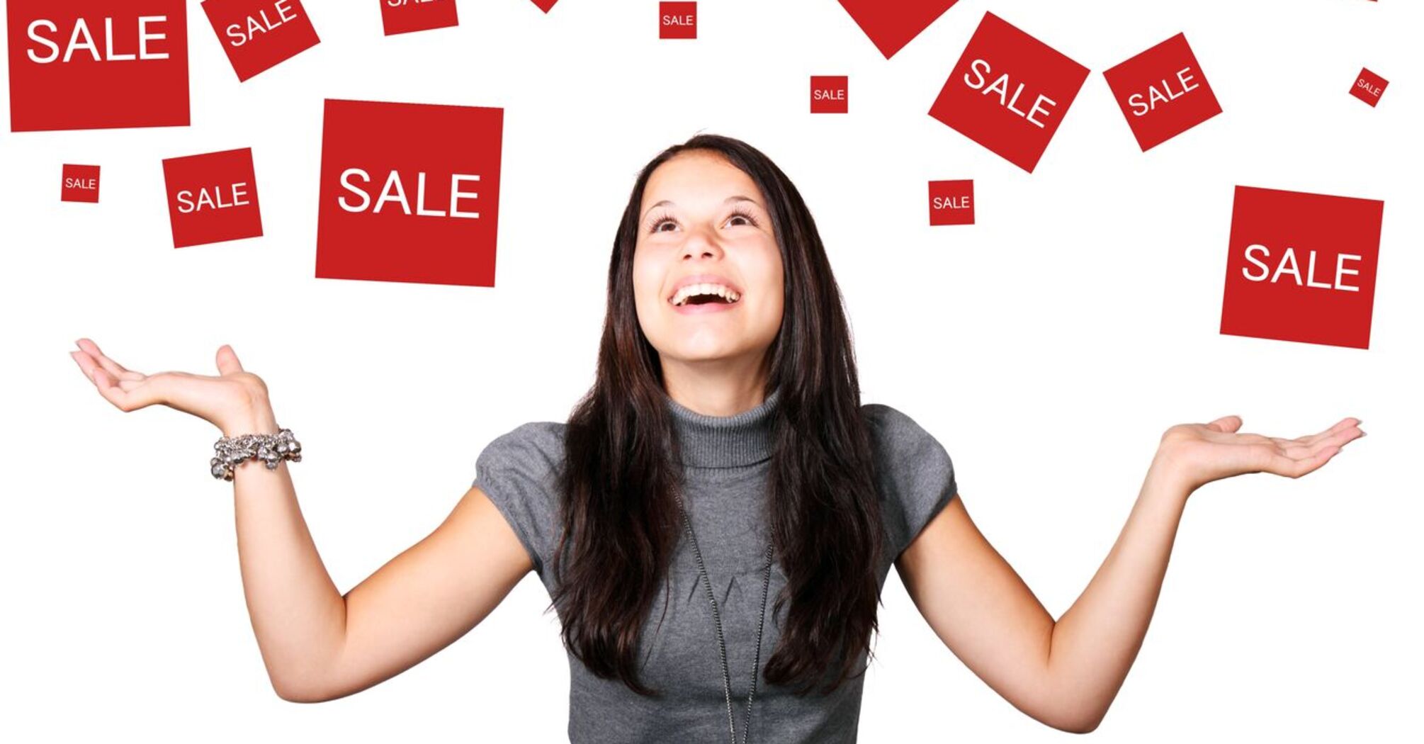 A woman smiling and looking upwards with open hands, surrounded by multiple red 'SALE' signs floating in the air against a white background 