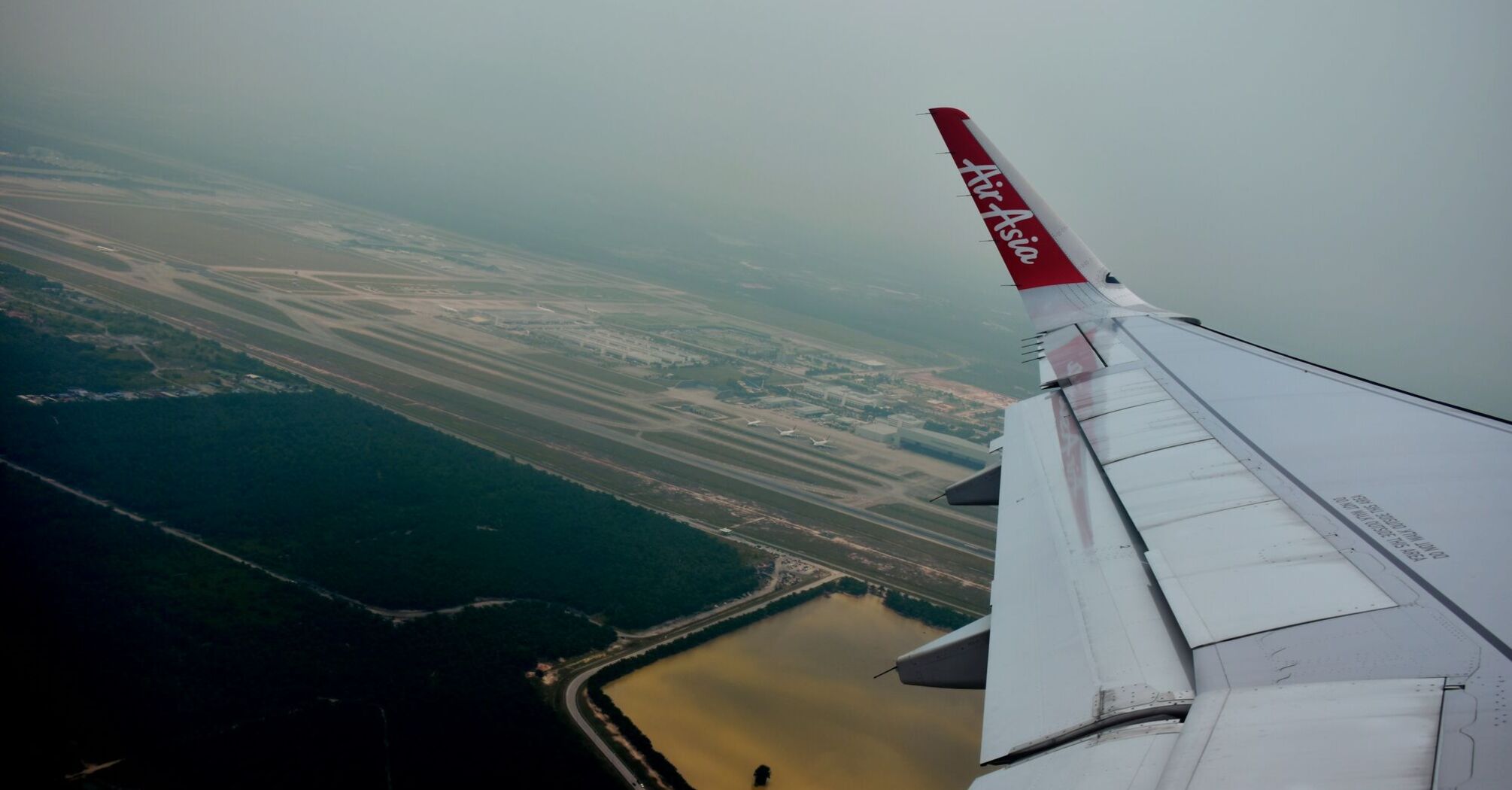 View from an airplane window showing the wing of an AirAsia aircraft with the airline's logo, flying over an airport runway and adjacent water body
