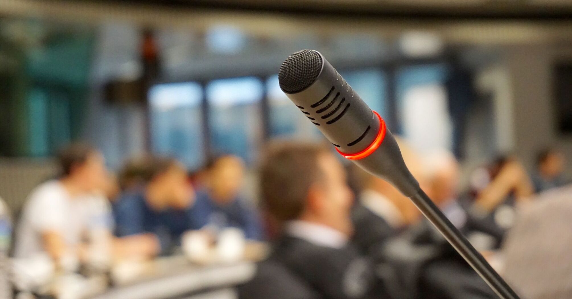 A microphone in focus with a glowing red light, against a blurred background of people in a conference or meeting setting
