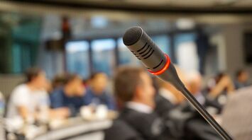 A microphone in focus with a glowing red light, against a blurred background of people in a conference or meeting setting