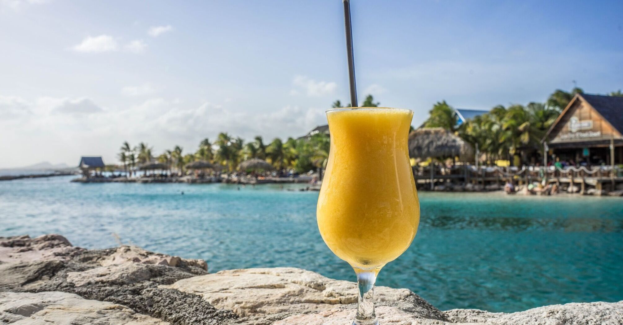 A passion fruit daiquiri in a tall glass with a straw, set on a rock with a blurred tropical beach resort background in Curacao