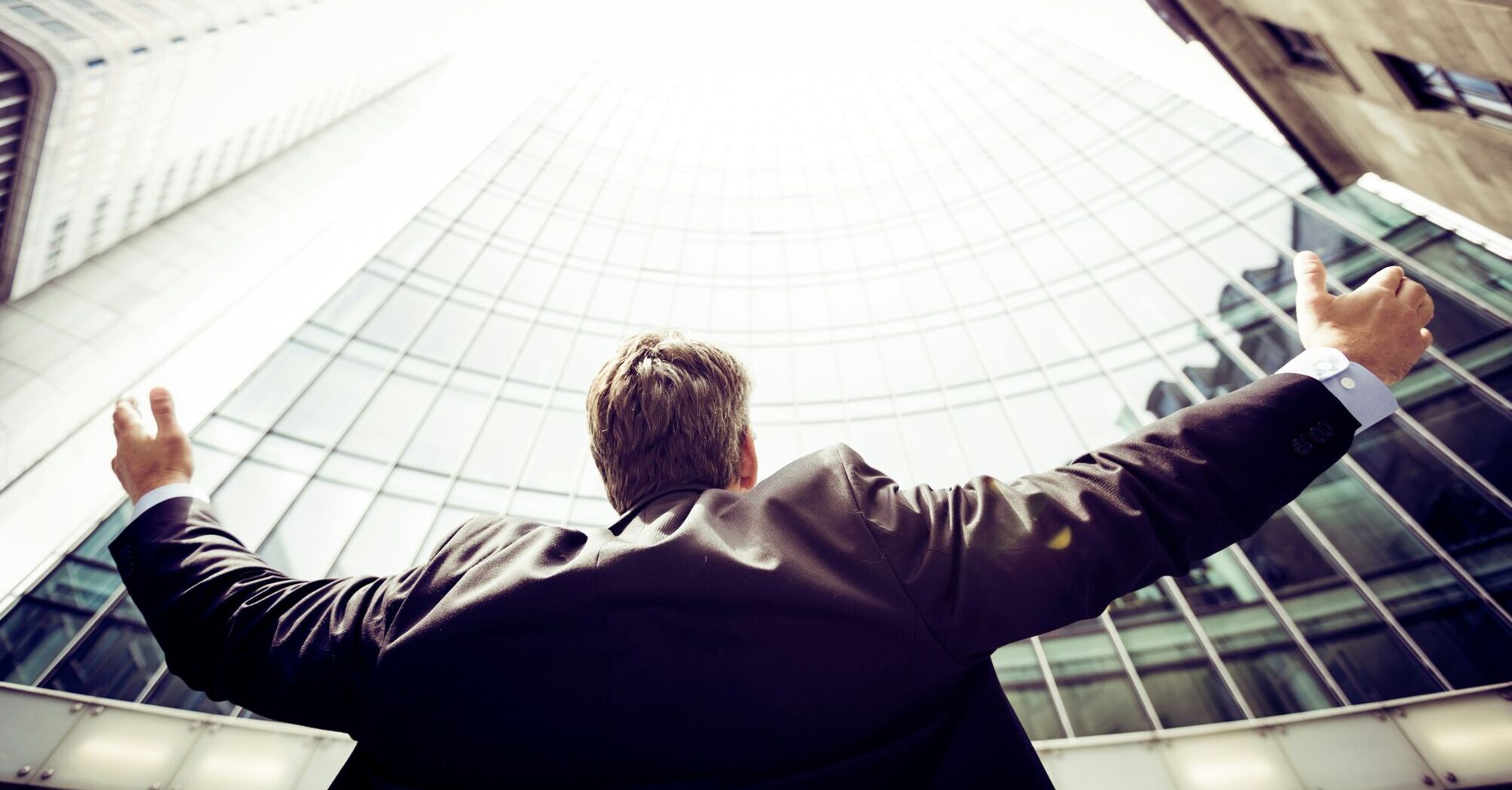 A man with outstretched arms facing a modern glass building, seemingly in a gesture of success or aspiration