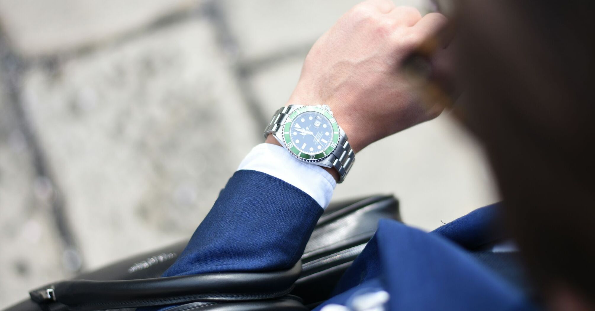 Close-up of a person's wrist wearing a luxury watch with a green bezel, white shirt, and blue suit