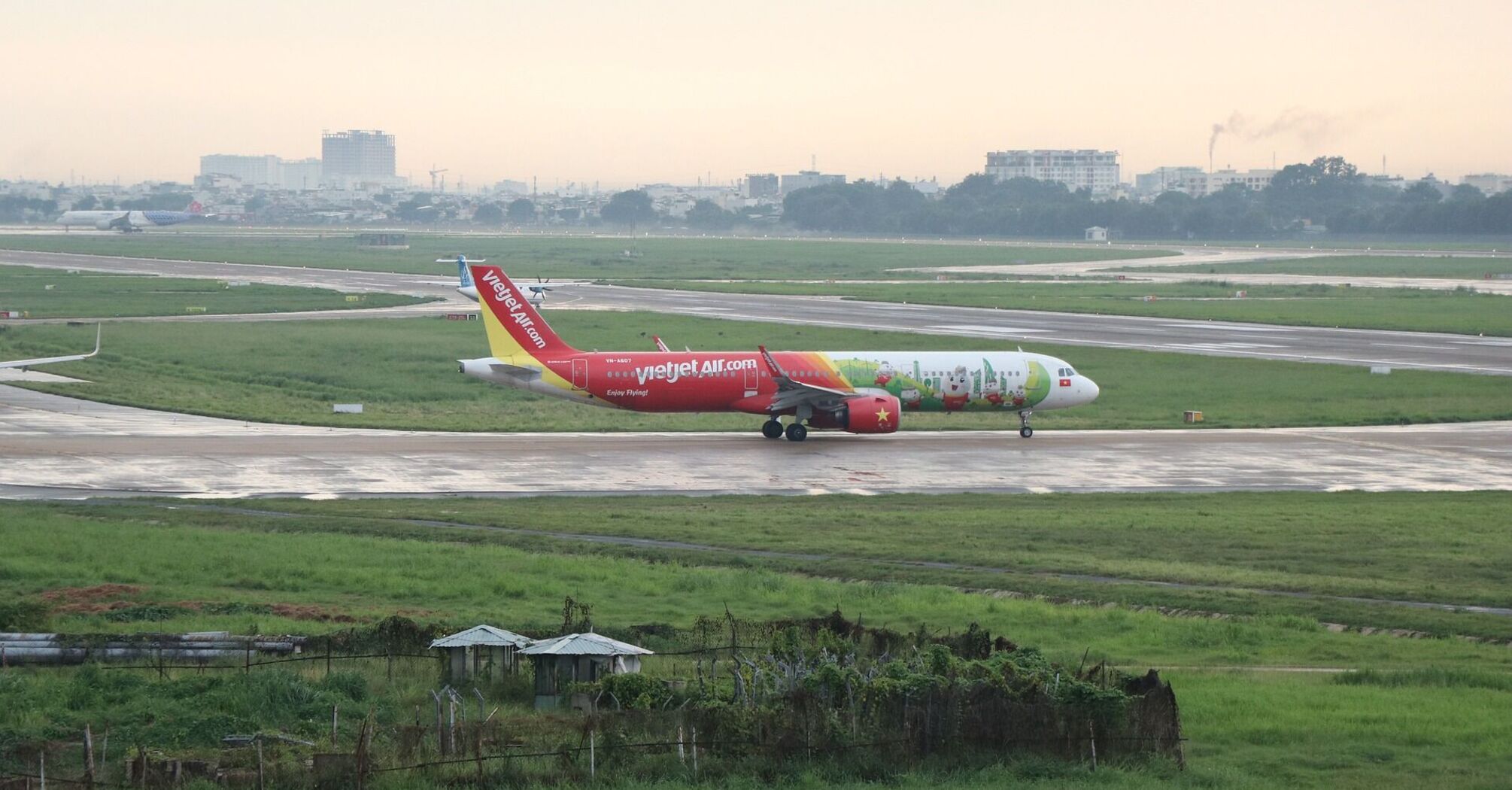 A Vietjet aircraft adorned with colorful decals, preparing for take-off on a runway with green fields and buildings in the background 