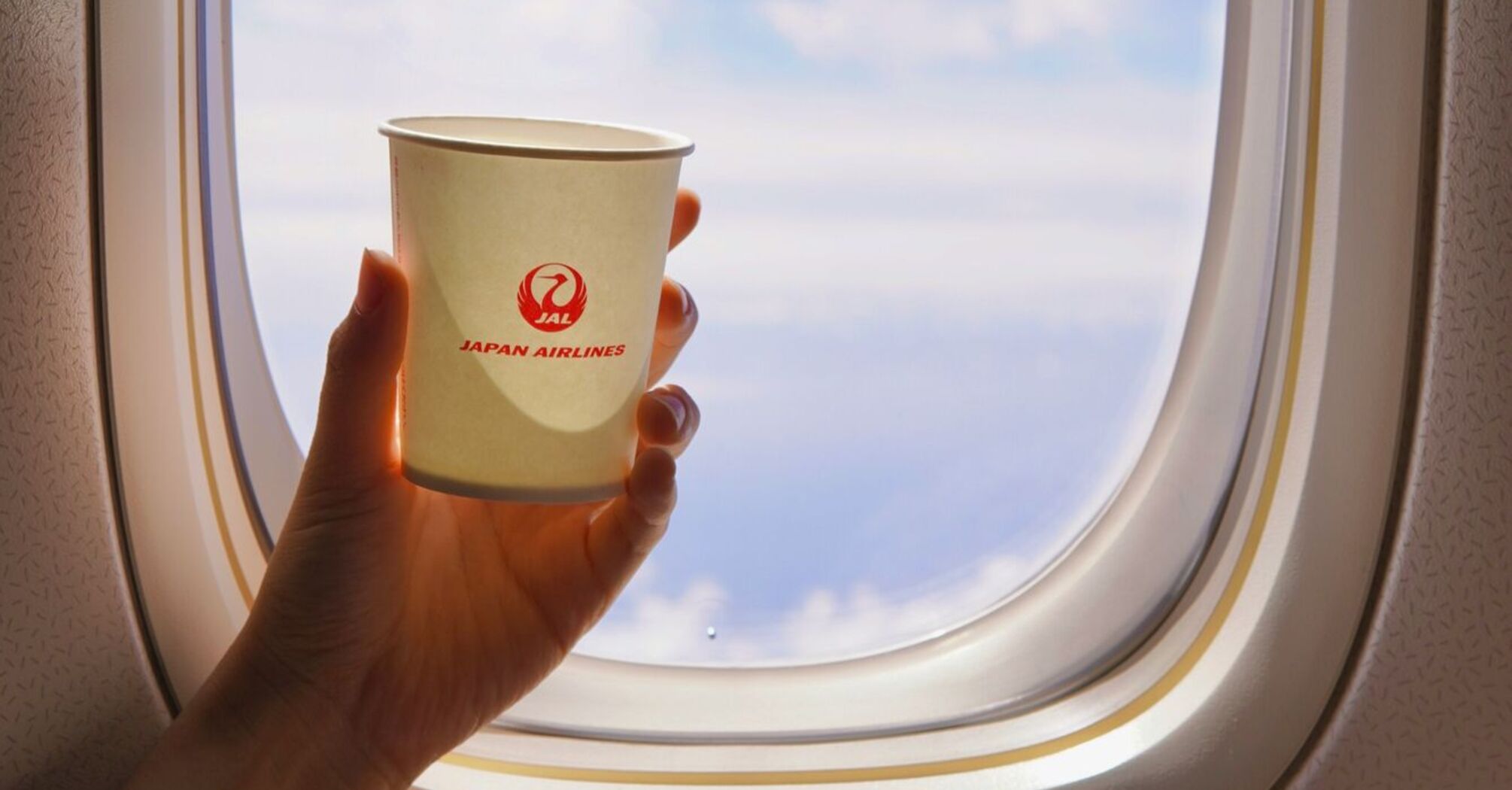 A hand holding a Japan Airlines branded paper cup against the background of an airplane window showing blue skies and clouds