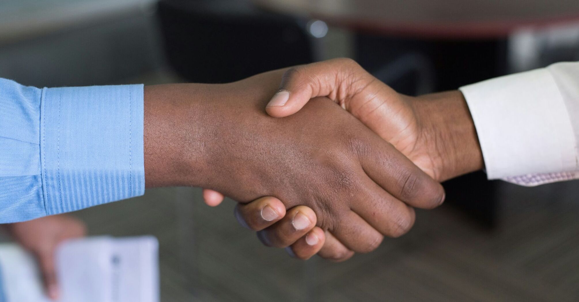 Two people shaking hands over a table in an office setting, symbolizing a partnership or agreement 