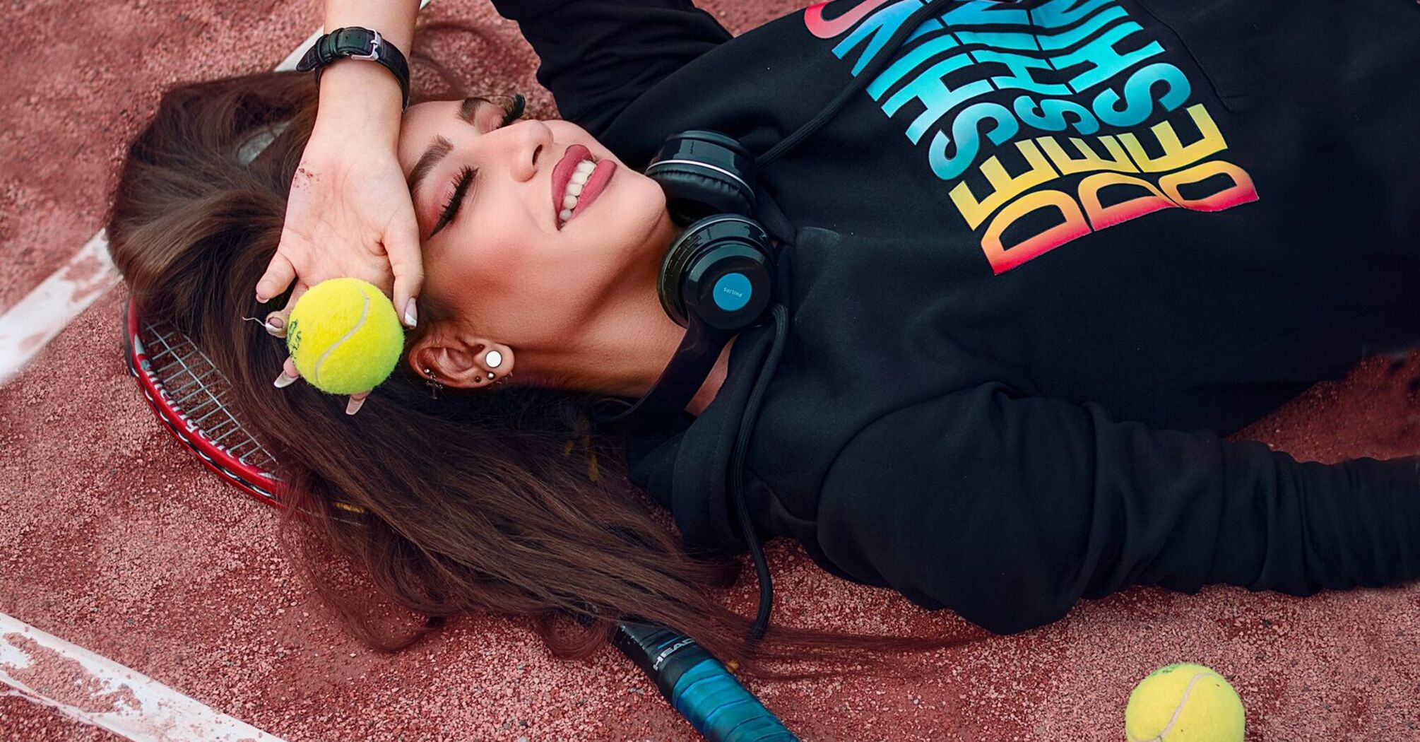 A woman lying on a clay tennis court with a tennis ball pressed against her forehead, a racket beside her, and headphones around her neck