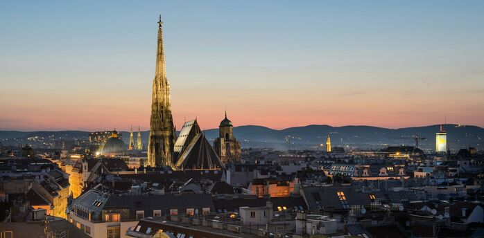 A cityscape at dusk with the skyline dominated by a cathedral's spire and other historical buildings, against a backdrop of mountains and a colorful sky.  Vienna, Austria