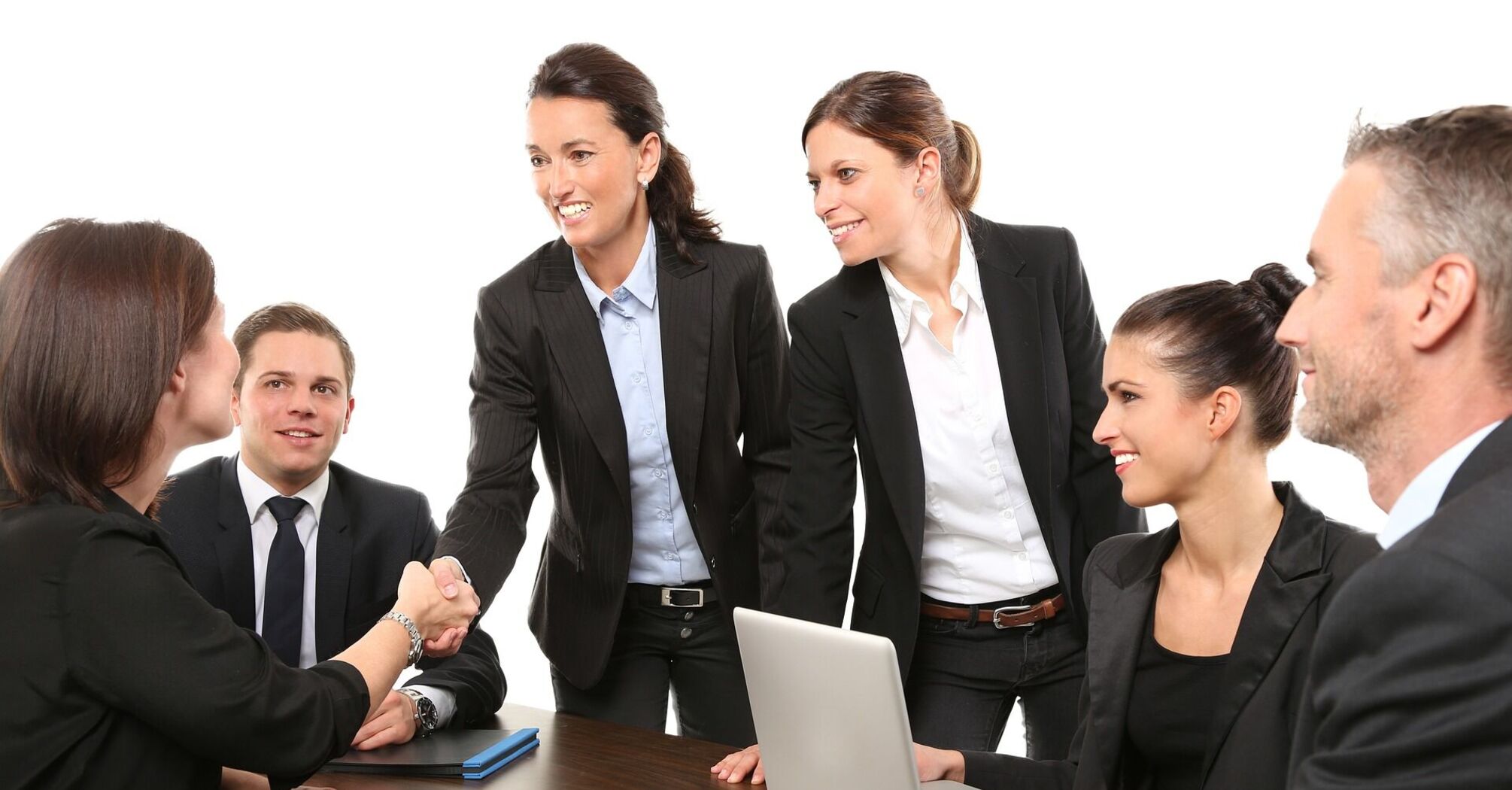 A group of professionals around a table with a laptop, engaging in a handshake and collaborative discussion
