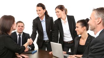 A group of professionals around a table with a laptop, engaging in a handshake and collaborative discussion