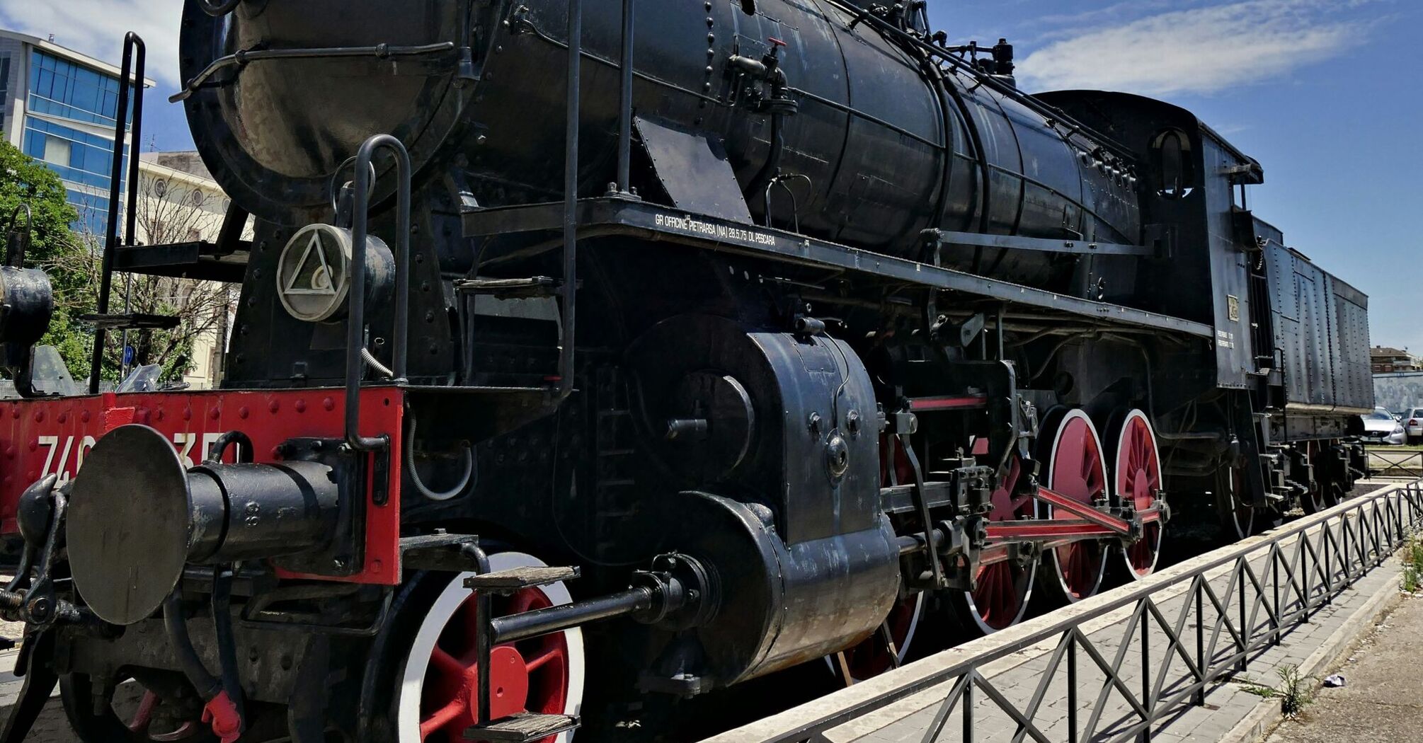 This is a photo of a vintage black steam locomotive with red wheels, displayed outdoors under a clear sky