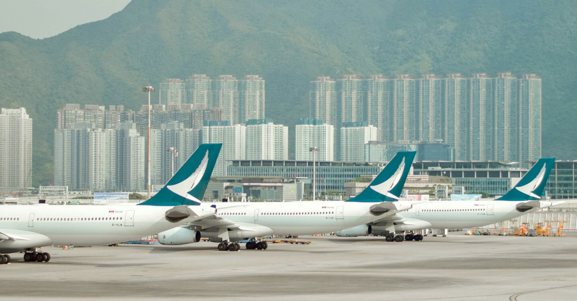 Cathay Pacific aircraft lined up at an airport with high-rise buildings and mountains in the background