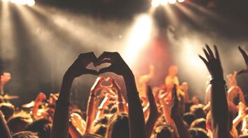 A crowd of concertgoers enjoying a live music performance, with many hands raised and one person making a heart shape with their hands against the backdrop of a brightly lit stage 