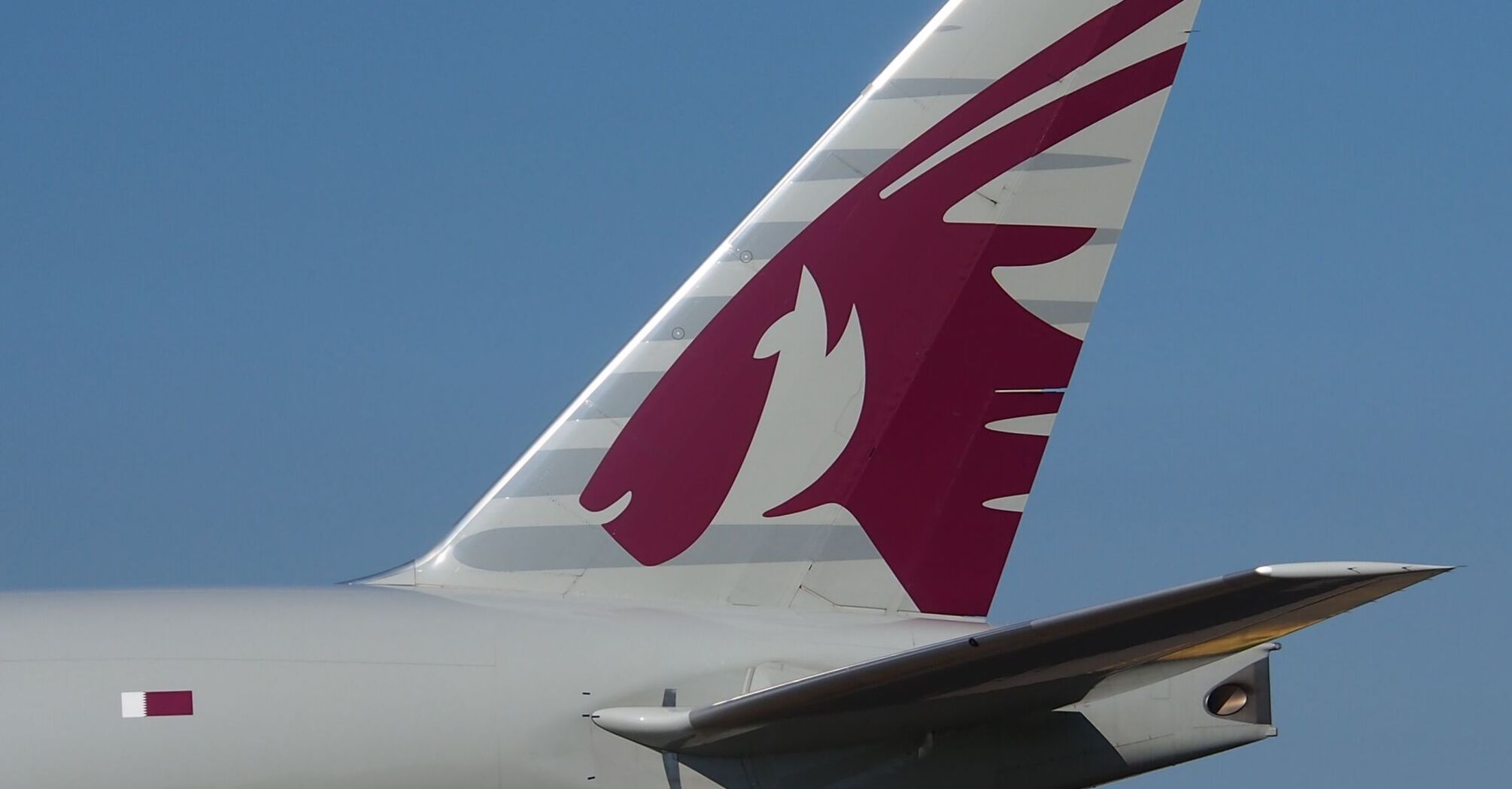 The tail fin of a Qatar Airways aircraft showing the airline's distinctive oryx logo against a clear blue sky 